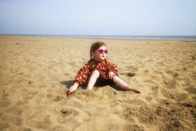 Image of a young girl in a leopard print dress and pink sunglasses sat burying her feet on a golden sandy beach. Behind her is the horizon, a clear blue sea and blue sky.