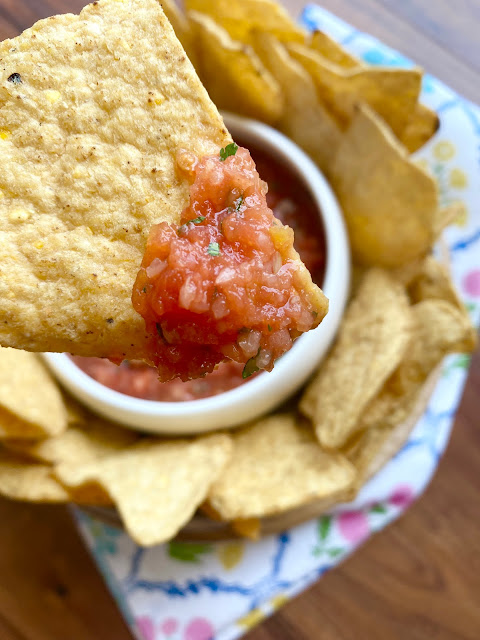 Close up of tortilla chip with salsa on it, with bowl of chips and salsa in the background.