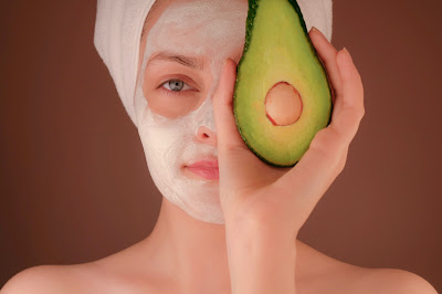 Mujer con mascarilla facial, el pelo envuelto en una toalla y sosteniendo medio aguacate