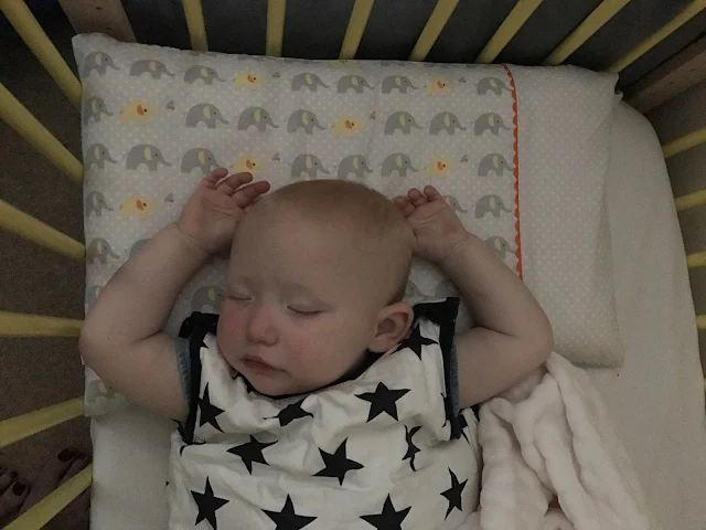 A 1 year old asleep in her cot with her arms above her head in a navy star sleeping bag on an elephant pillow