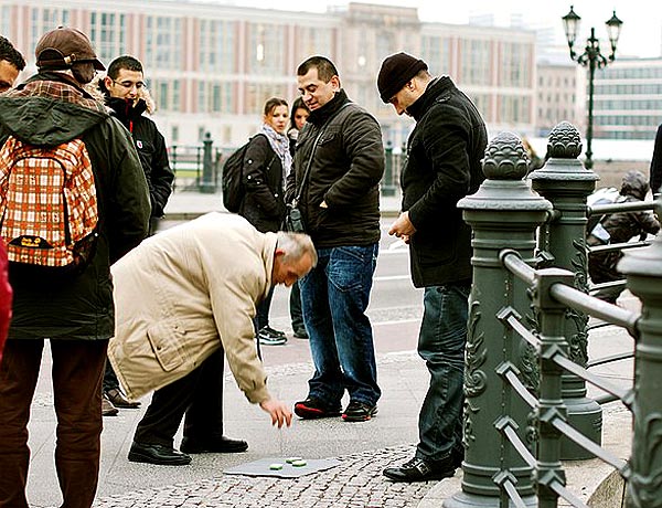 people on a sidewalk look on as an older man bends over to make his choice in a classic "shell game"