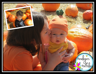 An older girl holding a baby girl and kissing her on the cheek, who is wearing a cute pumpkin hat, with pumpkins in the background