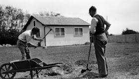 A black and white photograph of two men outside. One is digging at the ground with a pickaxe, while the other holds a shovel.