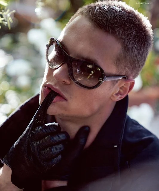 Hit model dude with short blonde hair sporting stylish sunglasses whine pulling off black leather gloves with his mouth viewfrom the neck up