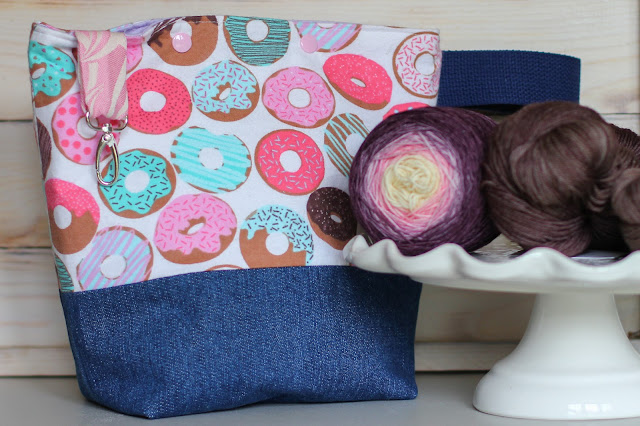 Donut print project bag with a denim boxed base is next to a cake stand that has a purple pink yellow gradient yarn cake next to brown yarn hanks. Project bag has a navy wrist loop on the side. A cotton candy pink and ecru fabric loop hangs over the top with a swivel silver snaphook.