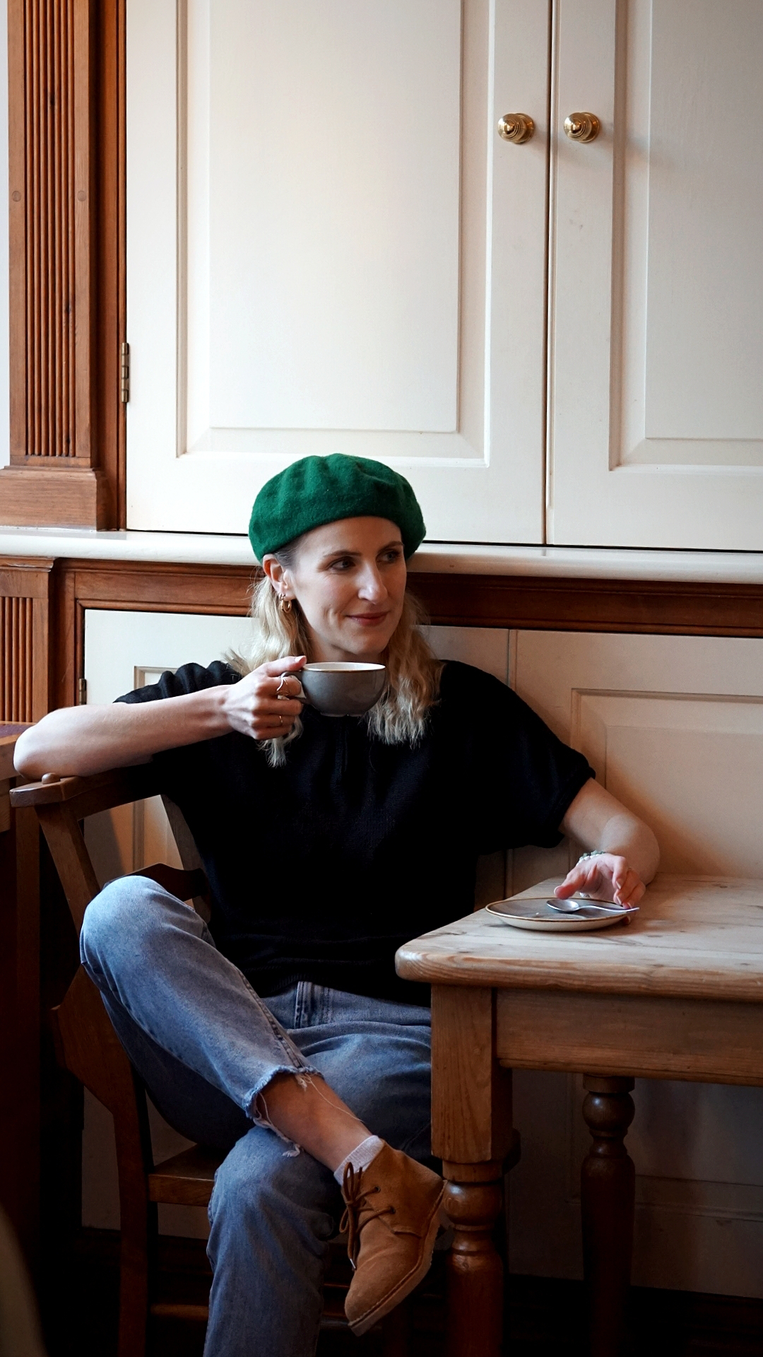 Amy is sat in a coffee shop drinking coffee. She is wearing a black short sleeved jumper, light blue jeans and a green beret.