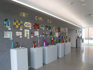 smooth white pillars and a gray wall display brightly colored children's artwork and the words "Art Camp 2019"