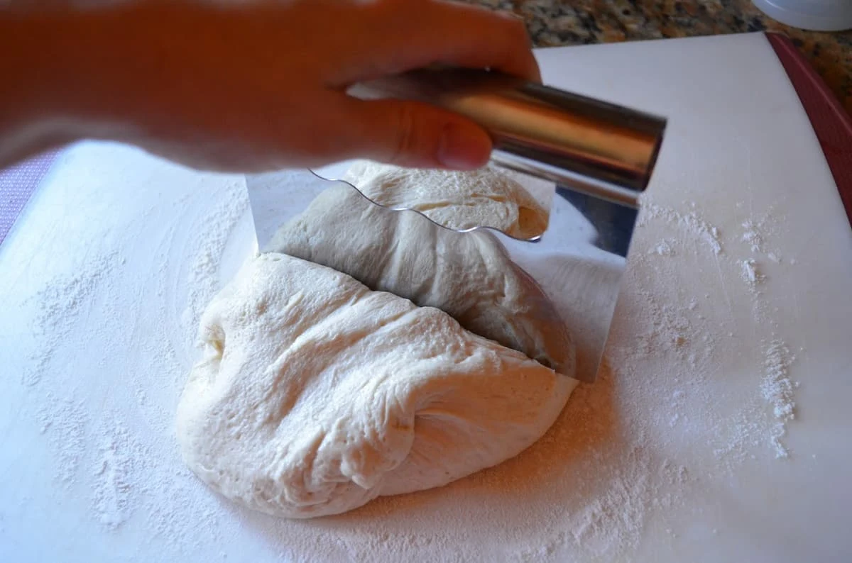 French Bread Dinner Roll dough on a white cutting board being divided into two with a bench scraper.