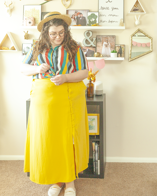 An outfit consisting of a straw boater hat, a light blue, yellow, green, white and black vertical striped collared short sleeve blouse with the ends tied into a bow on top of a yellow button down maxi skirt and white mules.