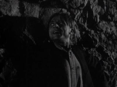 Dwight Frye in The Vampire Bat (1933) standing back to a cave wall, looking backward toward his pursuers.