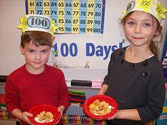 This is a picture of a boy and girl in a second grade class. They are holding a bowl with chips and wearing a hat that says one hundred. This picture was taken on the hundredth day of school party.