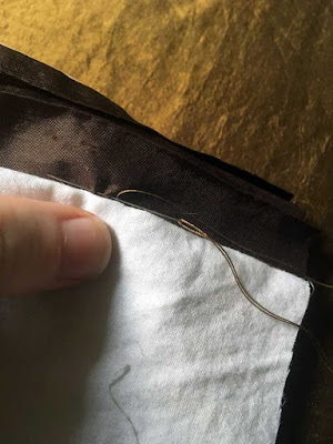 A close-up of a needle working a tiny running stitch in brown silk, just at the edge of folded-back white fabric, over a piece of brassy gold fabric.