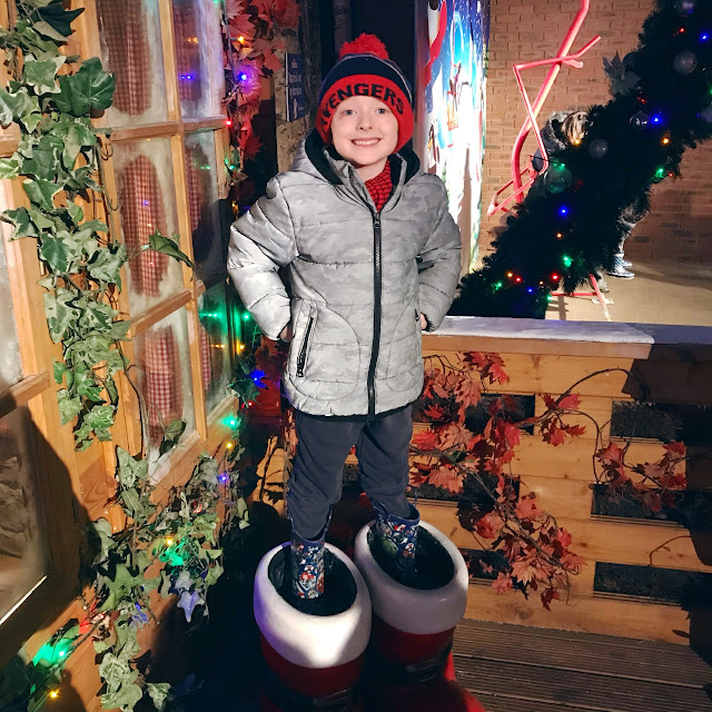 Little boy standing in a large pair of santas boots in front of a festive looking house