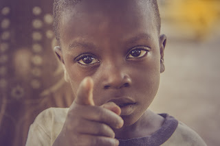 A photograph depicts a depressed young African boy extending his index finger at the foreground.