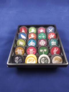 A tray containing the different tokens: black tokens with a Celtic knot raven, yellow tokens with a Celtic knot wolf head, deep yellow tokens with pikeman holding a shield, magenta tokens with a castle tower and spear, dark green tokens with two crossed battle axes, crimson tokens with a Greek warrior, olive tokens with a banner, bright blue tokens with a knight carrying sword and shield, deep purple tokens with a Celtic cross, teal tokens with warriors carrying spear and shield, deep blue tokens with a hawk, amber tokens with a saddled horse, bright green tokens with a bow and arrow, bright red tokens with a soldier mounted on horseback carrying a lance, burgundy tokens with a dagger piercing a bag of coins, lime green tokens with a rearing horse, reddish violet tokens with a crossbow, and dark blue tokens with two crossed swords.