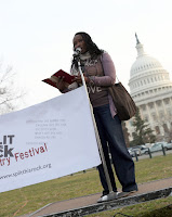 Image of a Black woman with braided hair at a microphone reading a poem from a book on a stage in front of the Capitol. To her left is part of a banner for Split This Rock Poetry Festival.