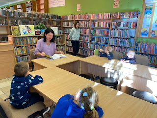 Pani bibliotekarka siedzi przed grupą dzieci i czyta im bajkę. Tło: regały z książkami i pani bibliotekarka.