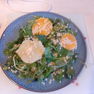 A blue dinner plate with giant cous cous, slices of grapefruit and orange, and green leaves on it.