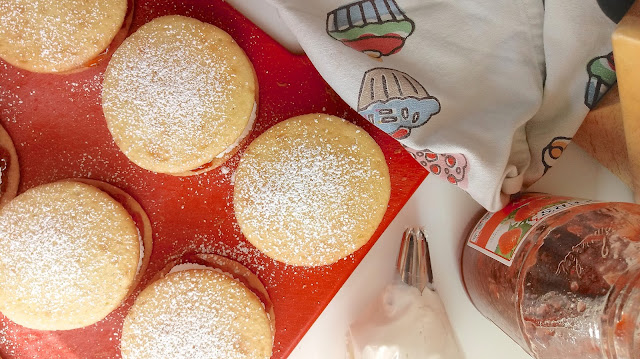 powdered sugar dusted finished Victoria Sandwich Whoopie Pies sitting on a red cutting board next to a tea towel, a jar of jam, and a piping bag full of frosting