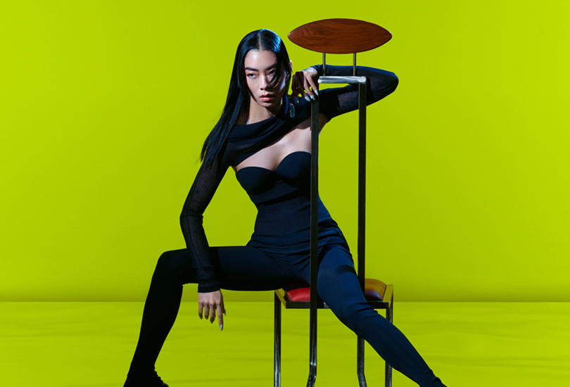 Rina Sawayama sat on a chair, in front of a neon green backdrop. Rina is wearing strapless black body suit, with a sheer black body-less top which covers part of her décolletage and arms.