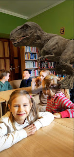 tło: sala biblioteczna. Dzieci siedzą przy stolikach. Na pierwszym planie dziewczynka w jasnym sweterku obok niej wygenerowany ze smartfona obrazek dinozaura tyranozaura. W tle regały z książkami.