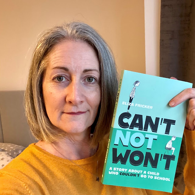 me, grey haired 50-year old in a yellow top, holding up Eliza Fricker's Can't Not Won't book. It has a green cover and an illustration of a mum at the top looking down on her child sitting on a school chair