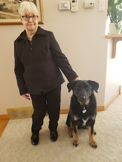 A small white-haired woman (Grandma Paulie) stands awkwardly beside Monty who is sitting on the carpet while Grandma Paulie gives him a pat on the head and poses for a photo.