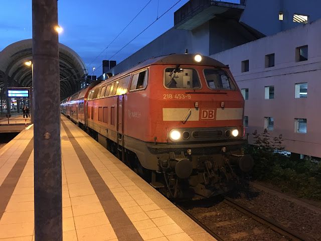 Von vorn schauen wir auf die BR 218 deren drei Frontlicher an sind und deren Führerstand erleuchtet ist. Gut ist die Typennummer 218 453-9 und das rote DB zu erkennen. Im Hintergrund ist ein Bogen der Kuppel des Hauptbahnhof Kiel zu sehen. Rechts neben der Lok eine weiße Mauer mit rechteckigen Löchern. Das ist ein Parkhaus.