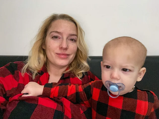 A tired looking mum (me) sitting up in bed in red and black chequed pyjamas with a baby boy sitting next to her in matching pyjamas