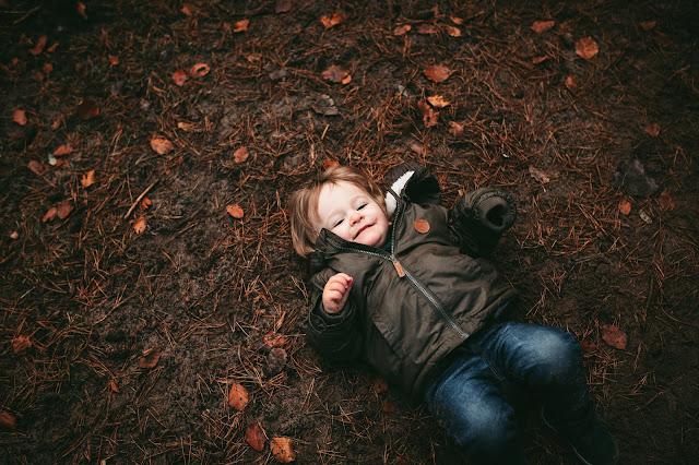 image shows a toddler boy lying on his back in the woods and looking to camera