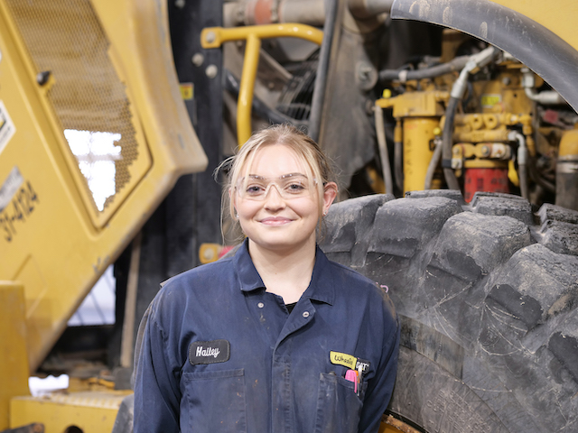 Hailey standing next to a tire of a diesel machine which towers her