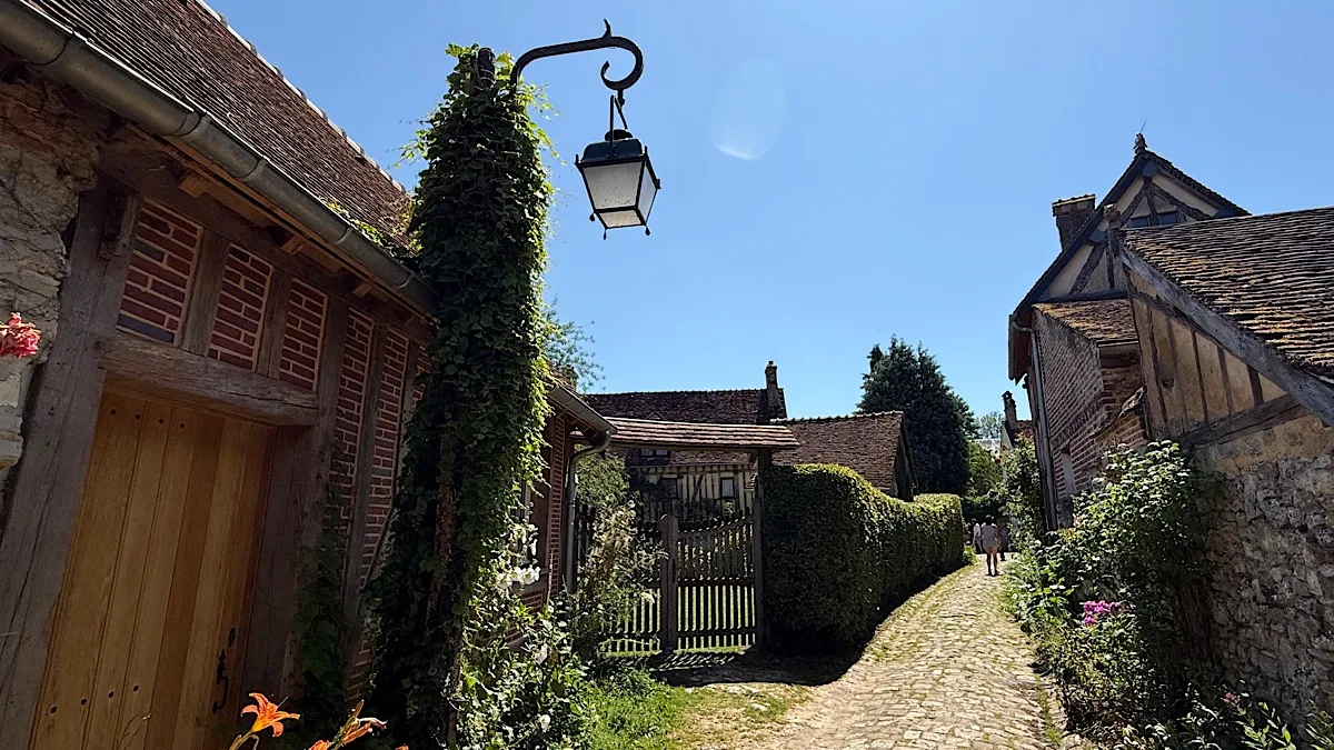 A country lane leads past some houses. On the left is a street lamp, gate and wooden door. On the right is the stone walls of another house.