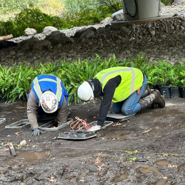 In the photo, two people are wearing bright blue and yellow construction vests and gear on the dirt ground installing vegetation underneath the SR 520 bridge on Foster Island.