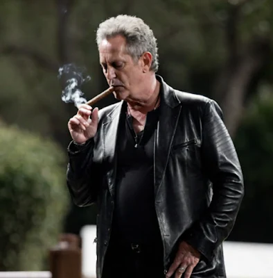 Lindsay Buckingham writing a cigar and wearing a black leather blazer standing outside from the knees up