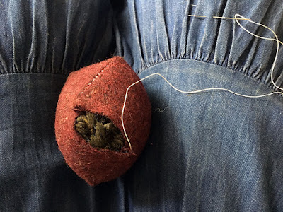 A red wool pincushion, shown from the side, with dark green yarn showing through an opening in the side seam. The pincushion is so firmly stuffed that the other seams are straining