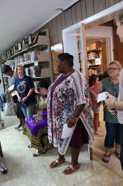 A woman addresses groups of people standing around the inside of a library.