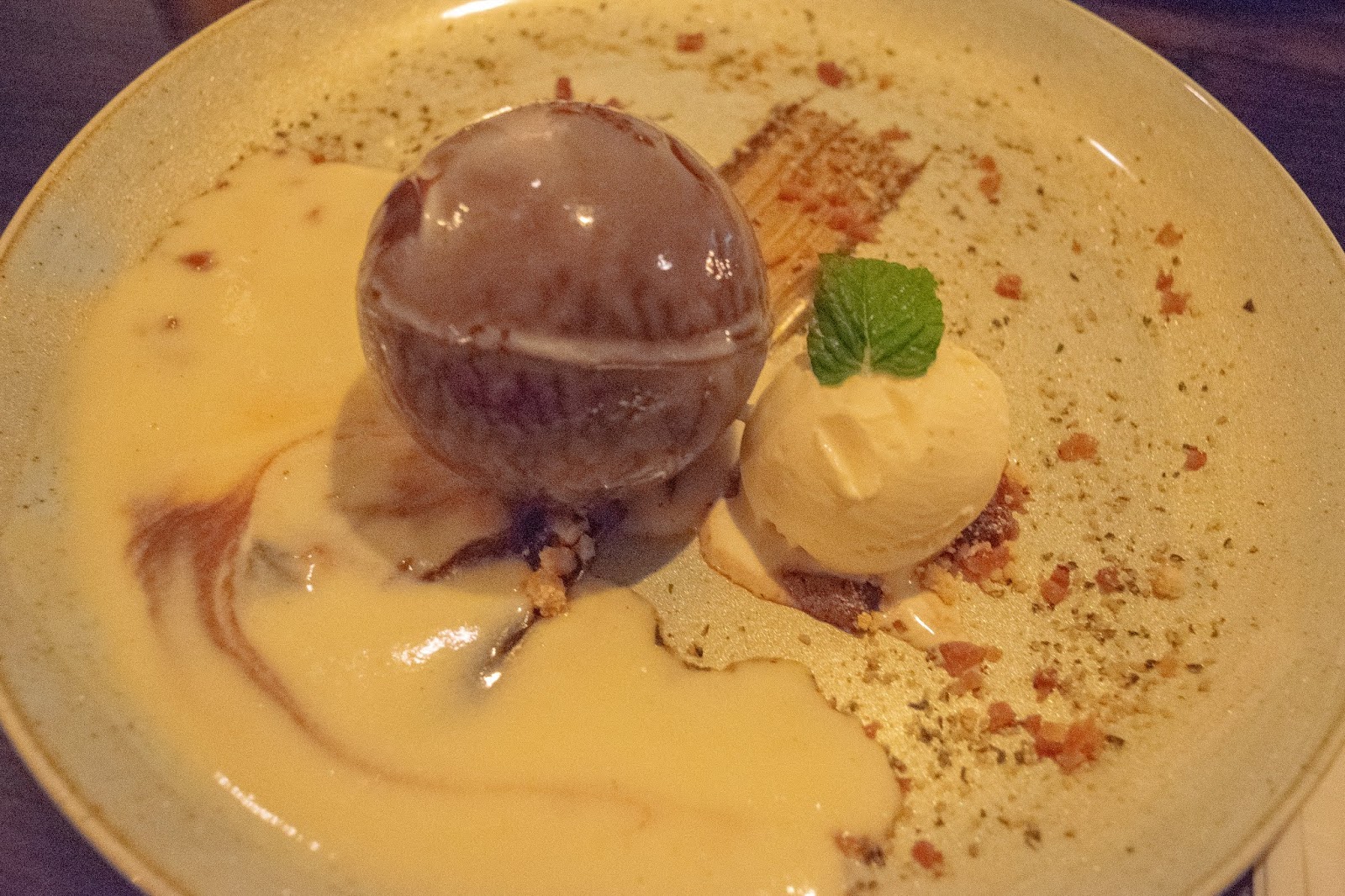 A chocolate sphere with white sauce over and a ball of vanilla icecream.