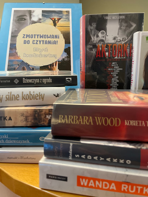 Na ladzie bibliotecznej znajduje się pionowy plakat "Zmotywowani do czytania: Być kobietą" na małej sztaludze oraz kilka książek o kobietach ("Aktorki", "Trzy silne kobiety" , "Dziewczyna z ogrodu" ).