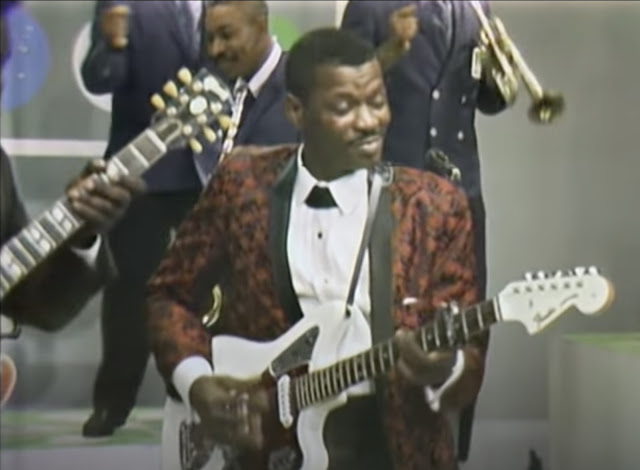 Screenshot of Clarence "Gatemouth" Brown playing on the TV show "The Beat" where he's smiling and playing his guitar. The band is in back of him and part of Freddy King's hand and guitar are visible in the shot. Brown is wearing a red brocade suit.