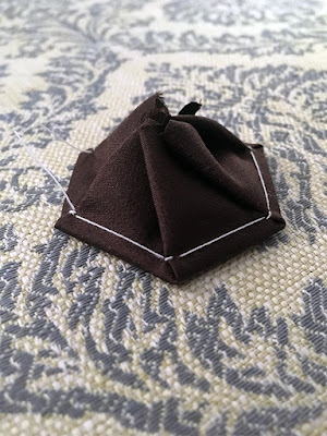 A close-up of a single silk hexagon, upside down on blue-and-white brocade fabric, with the seam allowance carefully arranged into a neat peak of pleats. The hexagon looks black. It's not. But apparently it wants to be.