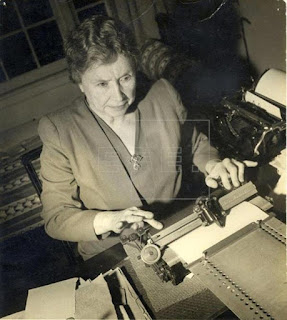 La foto en blanco y negro muestra a Hellen Keller, una señora de edad, rostro amable, pelo corto negro, vestido mangas largas color gris, tipea en una máquina de escribir Braille. Está sentada frente a una mesa de trabajo.