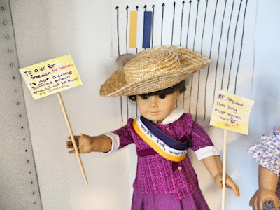 American Girl doll in plum colored tweed jacket and skirt, straw hat, holding a yellow and purple sign asking for freedom for female prisoners