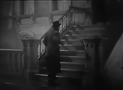 A shadowy figure in a trenchcoat and hat climbs the stairs outside a large terraced house