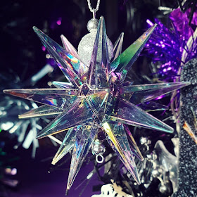 A bauble in the shape of a jaggedly pointed star with many points, made of clear plastic with an iridescent rainbow finish that tends towards green and purple. The bail for suspending the bauble is two silver beads It is infront of the black Christmas tree and a dark purple wall. Bright purple tinsel is visible 