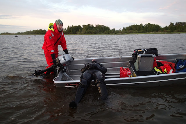 Sukeltaja makaa puoliksi veneessä, toinen ihminen työntää venettä matalassa rantavedessä. 