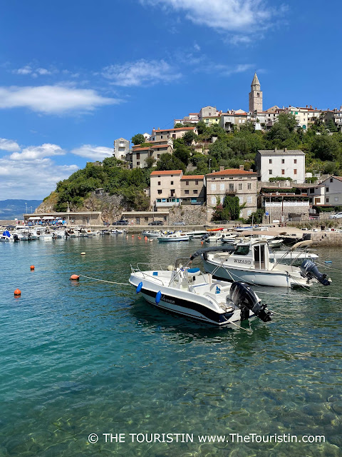 White middle-sized yachts in a harbour with crystal clear water at the foot of a small village with a stone church tower in its centre of red-roofed houses, under a bright blue sky flecked with only a few fluffy white clouds.