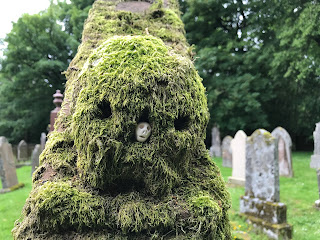 A close up photo of a small ceramic skull (Skulferatu 79) in the nasal cavity of the moss covered carved skull on David Scott's gravestone at Durisdeer.  Photograph taken by Kevin Nosferatu for the Skulferatu Project.