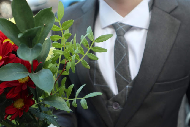 Flowers in the foreground, and a boy wearing a waistcoat and tie, with a contrasting grey jacket over the top slightly in the background. 