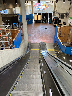 Photo from top of escalators of a tiled area surrounded by blue work walls. There is a painting over the fare machines.