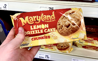 A white hand holding a rectangular packet containing light brown circular cookies with Maryland cookies lemon drizzle cake chunkiest in yellow and white font on a bright background